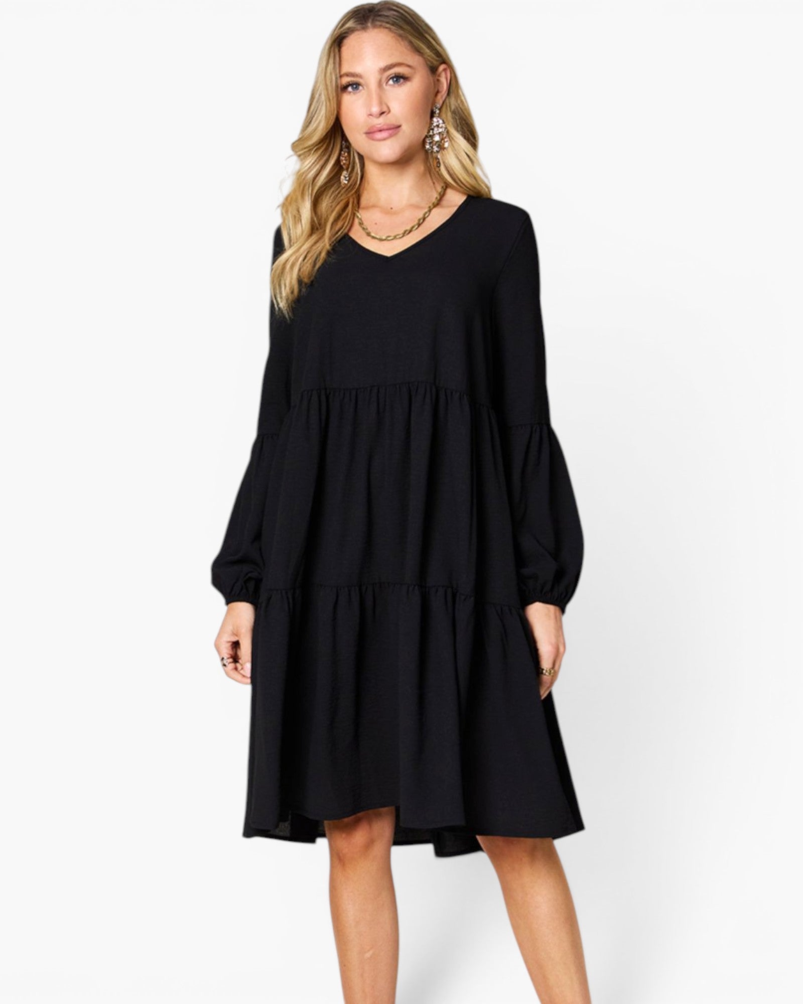 A woman modeling a black long-sleeve tiered dress with a V-neckline, photographed against a clean white studio background.