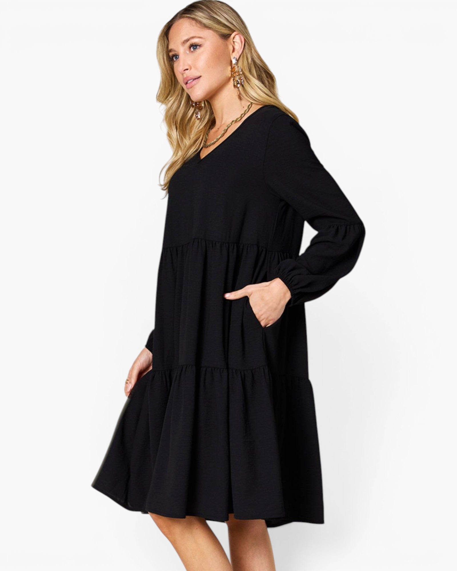 A woman modeling a black long-sleeve tiered dress with pockets, photographed against a plain white studio background.