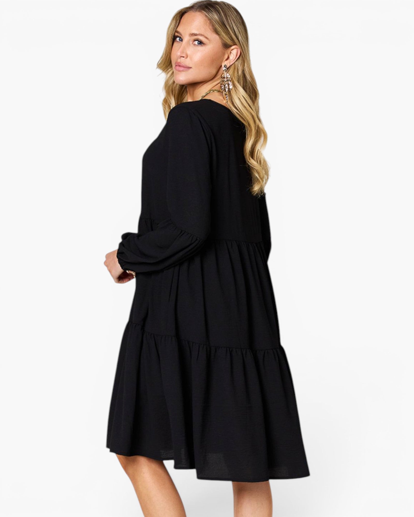 A woman modeling a black long-sleeve tiered dress, shown from a three-quarter back angle against a plain white studio background.