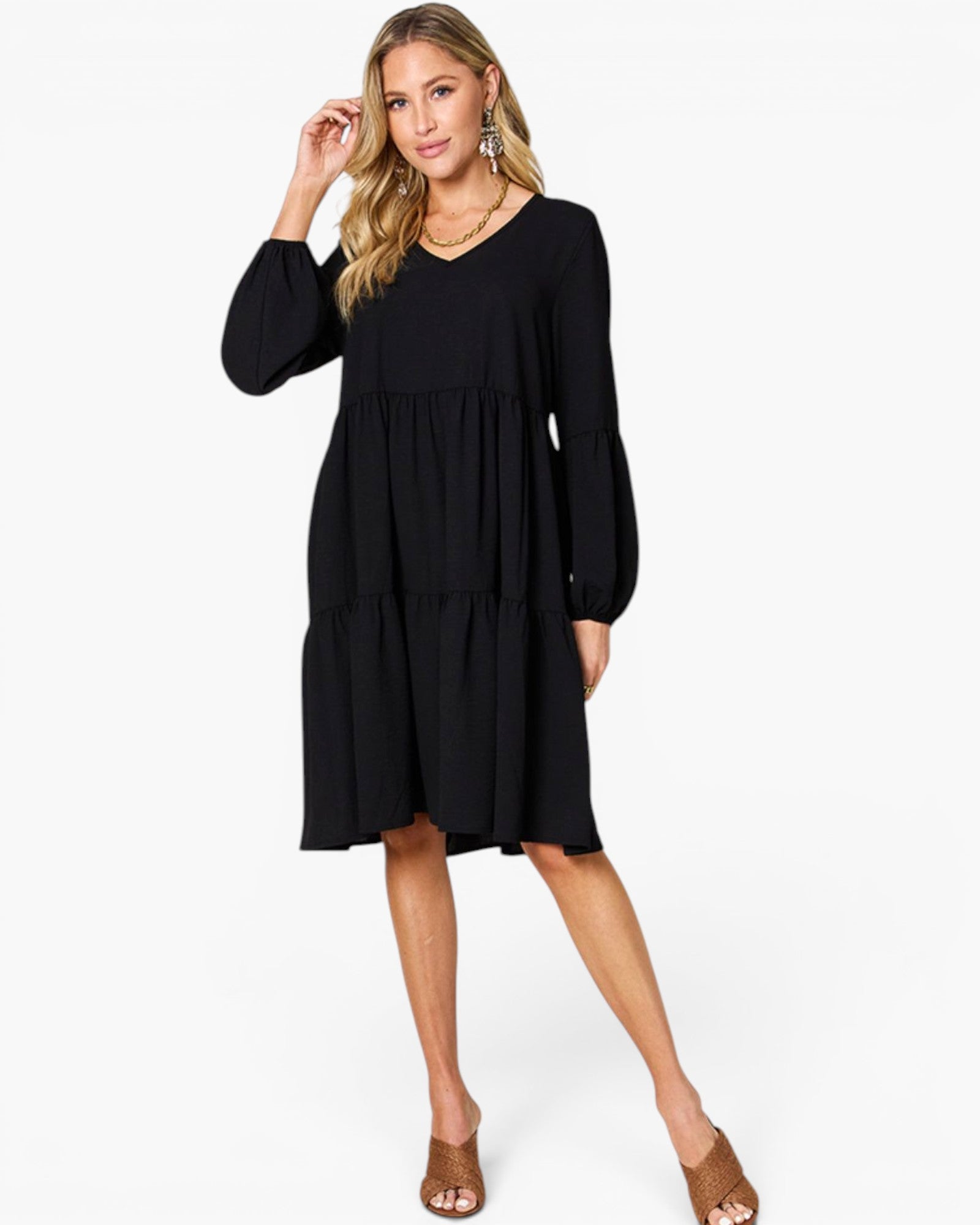 A woman modeling a black tiered long-sleeve dress with balloon sleeves, paired with brown mule sandals and gold jewelry, against a plain white studio background.