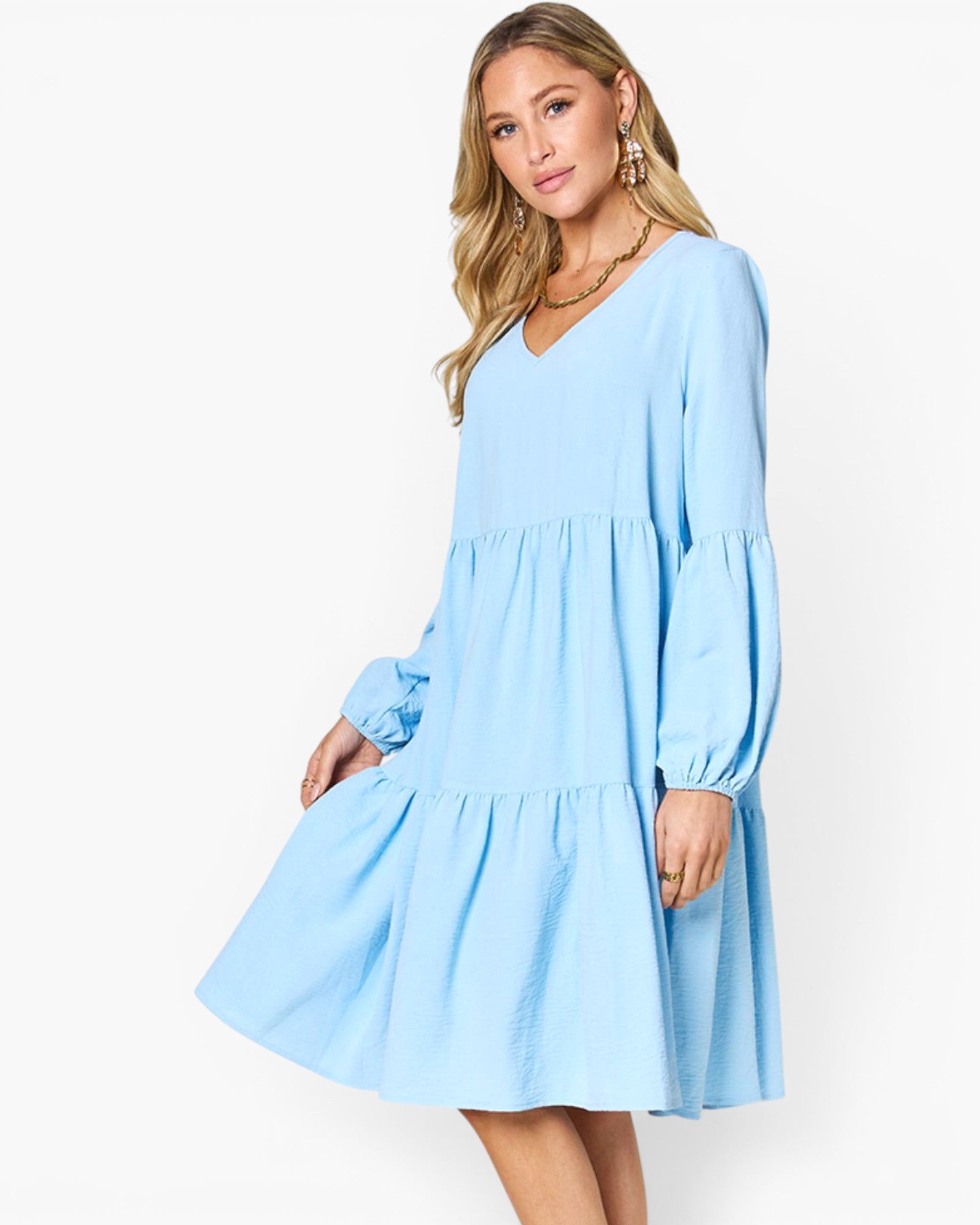 A woman modeling a light blue tiered long-sleeve dress with a V-neckline, photographed against a clean white studio background.