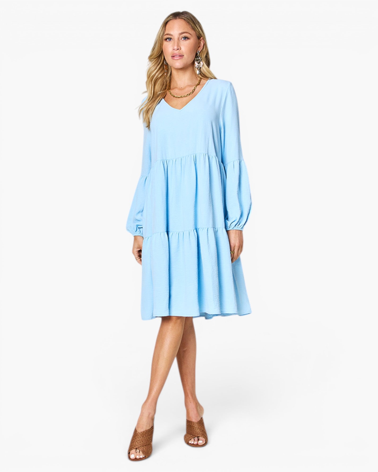 A woman modeling a light blue tiered long-sleeve dress with a V-neckline, paired with brown mule sandals, against a plain white studio background.