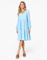A woman modeling a light blue tiered long-sleeve dress with a V-neckline, paired with brown mule sandals, against a plain white studio background.