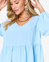 A woman wearing a light blue tiered V-neck dress with long puff sleeves, paired with gold statement earrings and a gold chain necklace, against a plain white background.