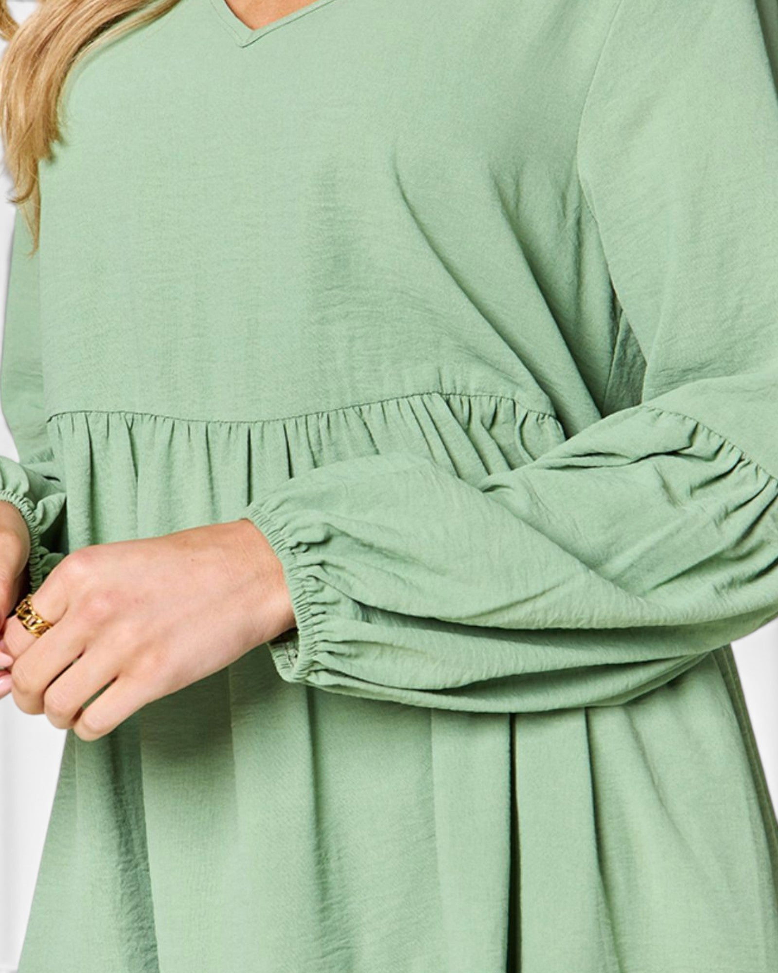 A close-up of a person wearing a sage green long-sleeve dress with a gathered waist and ruched sleeves, shown against a plain light background.