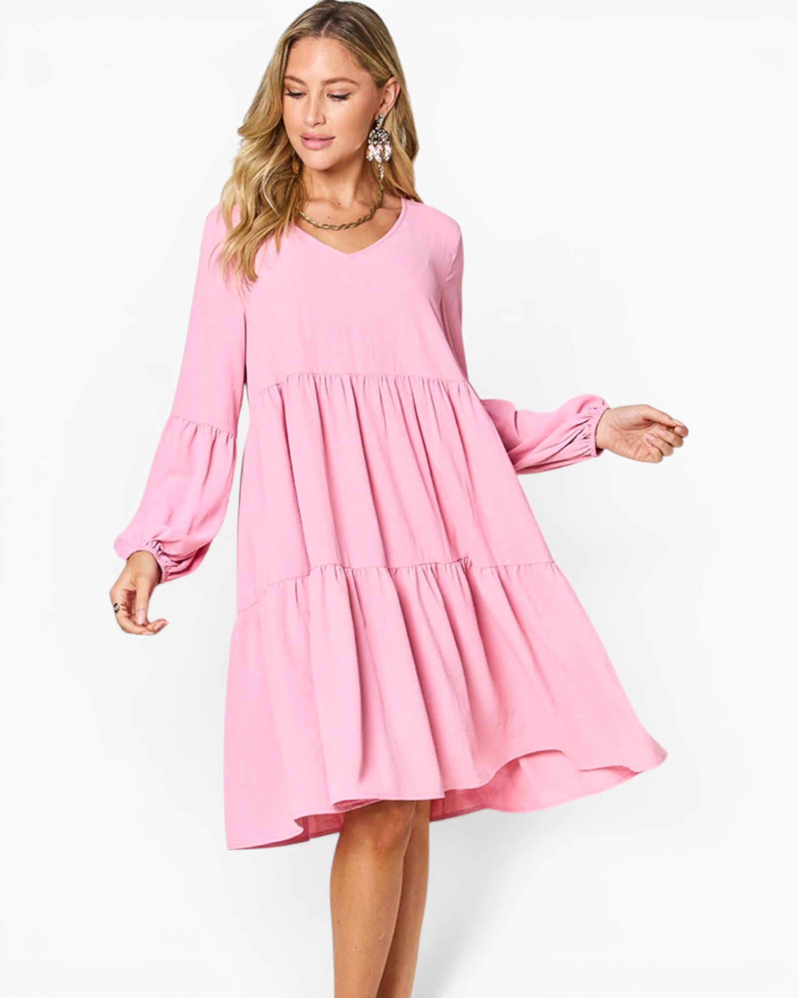 A woman modeling a pink tiered long-sleeve dress with a V-neckline against a plain white studio background.