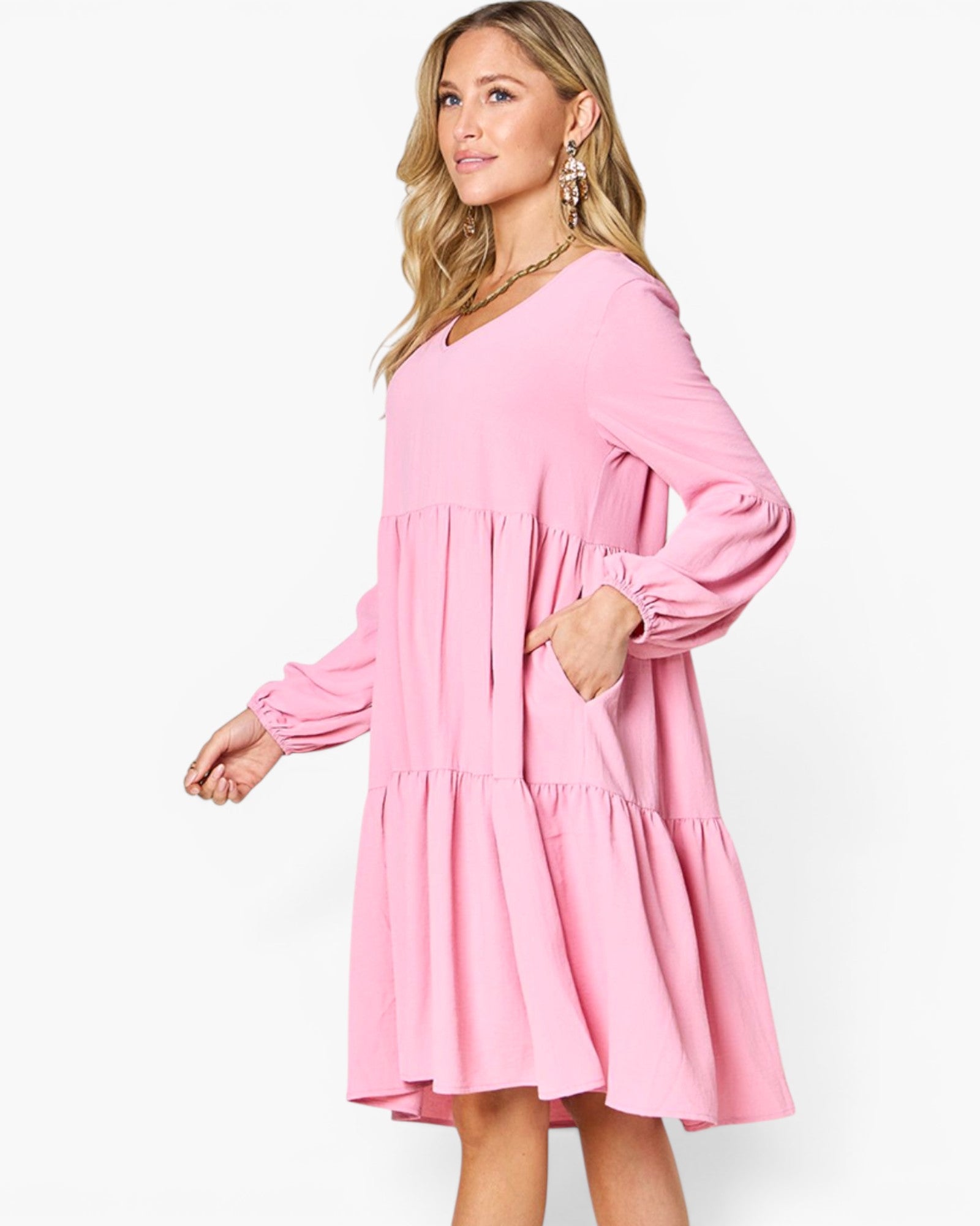 A woman modeling a pink tiered long-sleeve dress with a V-neckline and elbow-length sleeves, posed against a clean white studio background.