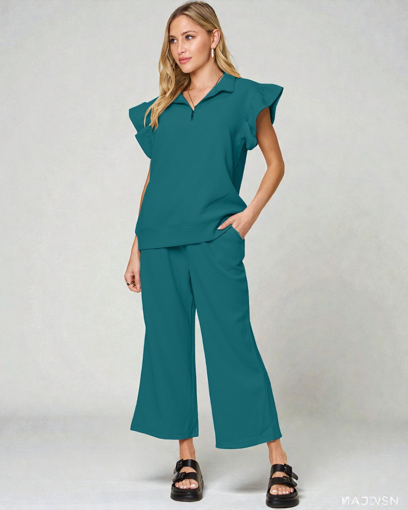 Woman wearing a teal outfit with ruffled top and wide-leg pants on a light gray background