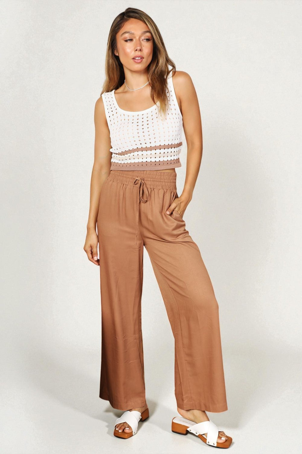 Woman wearing a white sleeveless top and brown wide-leg pants on a white background