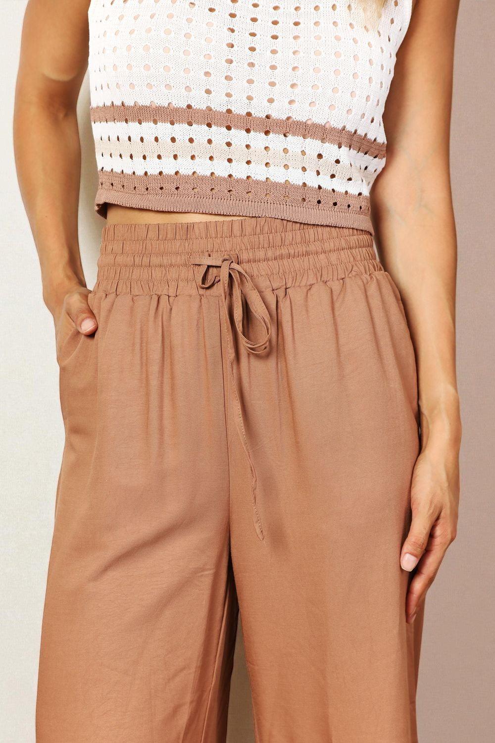 Person wearing a white top with brown accents and brown pants against a beige background