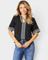 Woman wearing a black blouse with white embroidery and blue jeans on a plain background