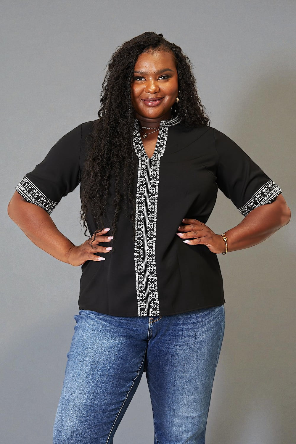 Woman wearing a black top with white embroidery and blue jeans against a gray background