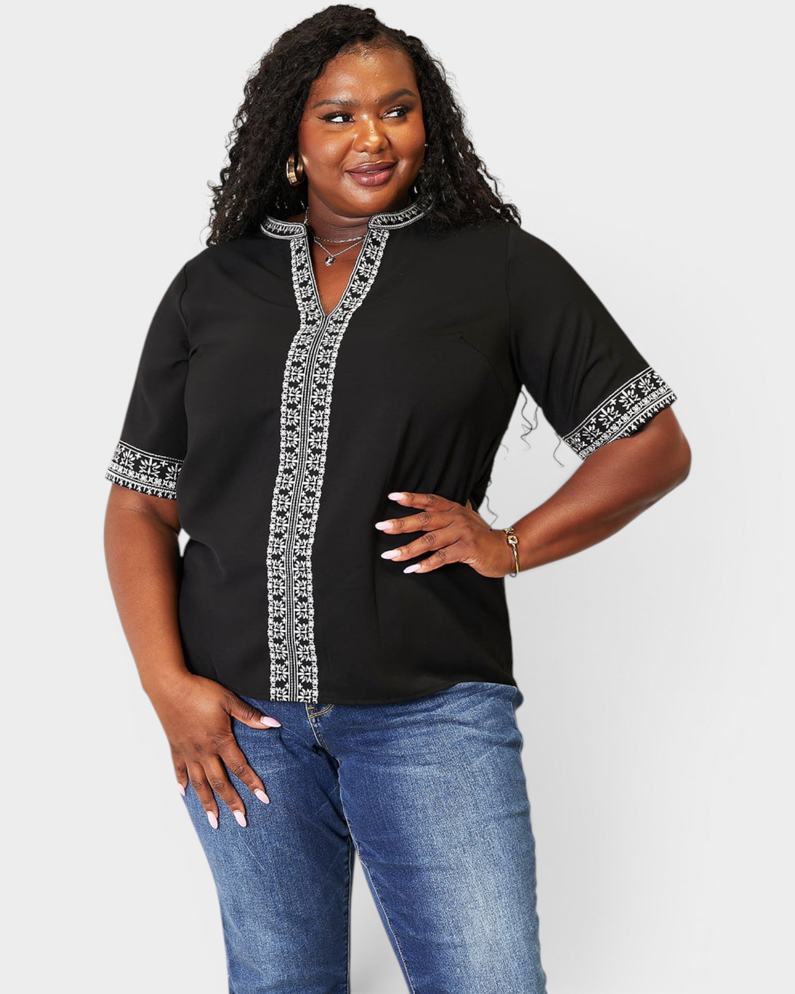 Woman wearing a black blouse with white embroidery and blue jeans on a light gray background