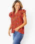 A woman wearing a red short-sleeve blouse with a black and orange floral print and ruffled shoulders, paired with blue jeans, against a light gray studio background.