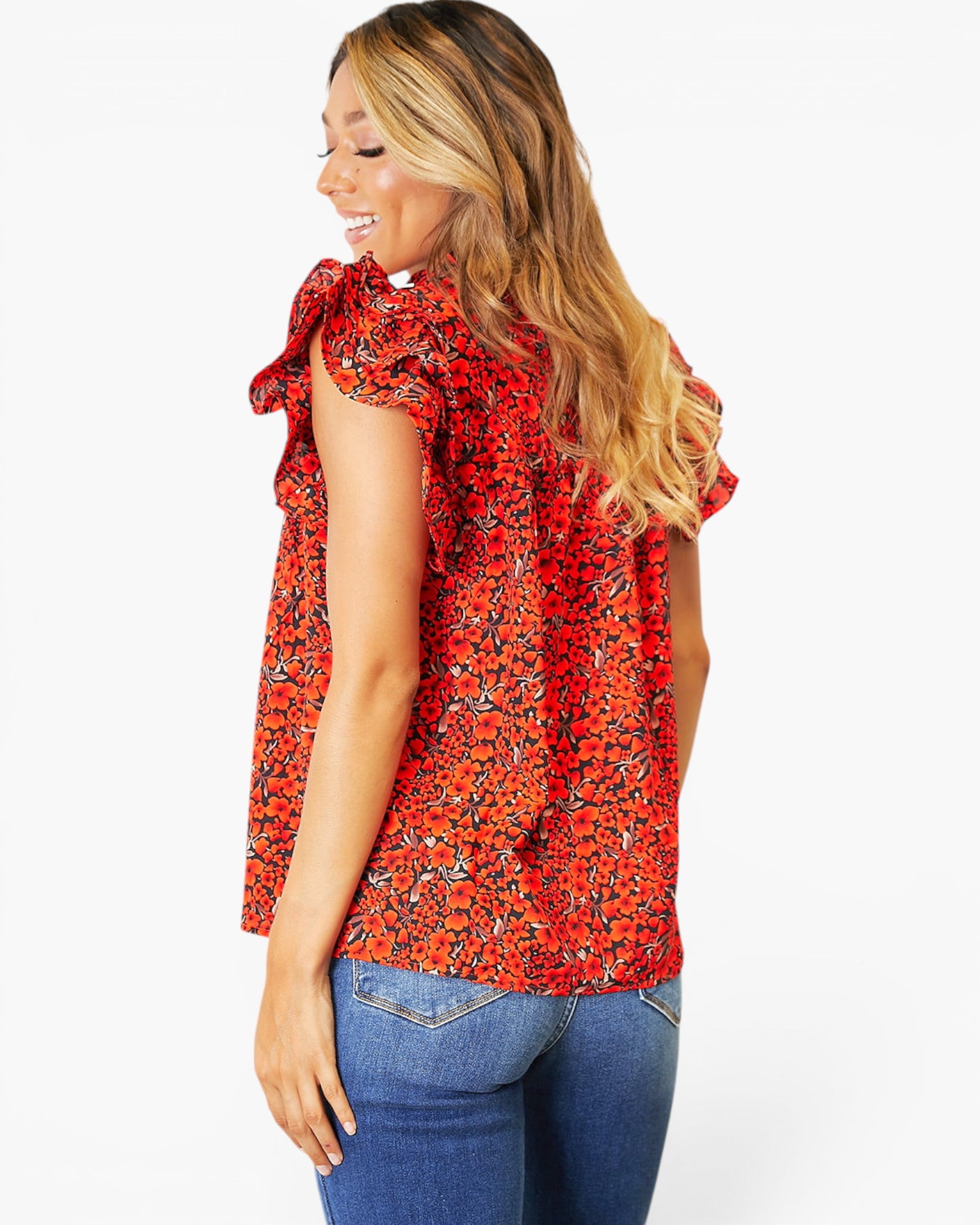 A woman shown from the back wearing a red sleeveless blouse with a dense black and orange floral print, ruffled shoulder details, and blue jeans, set against a plain white background.