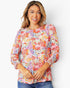 A woman wearing a long-sleeve blouse with a vibrant multicolor floral and abstract print, paired with blue jeans, photographed against a plain white studio background.