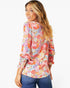 A woman wearing a colorful floral print long-sleeve blouse and blue jeans, photographed against a plain light gray studio background.