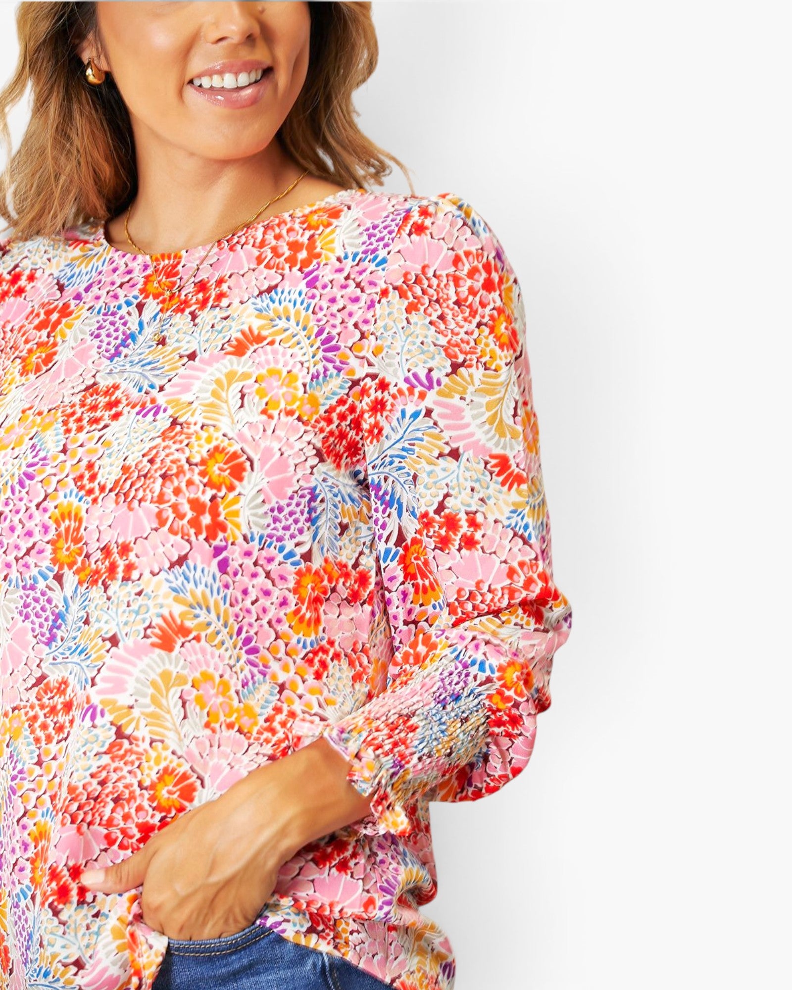 A woman wearing a long-sleeve blouse with a dense multicolor floral and abstract print on a light pink base, paired with blue jeans against a plain white background.