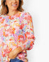 A woman wearing a long-sleeve blouse with a dense multicolor floral and abstract print on a light pink base, paired with blue jeans against a plain white background.
