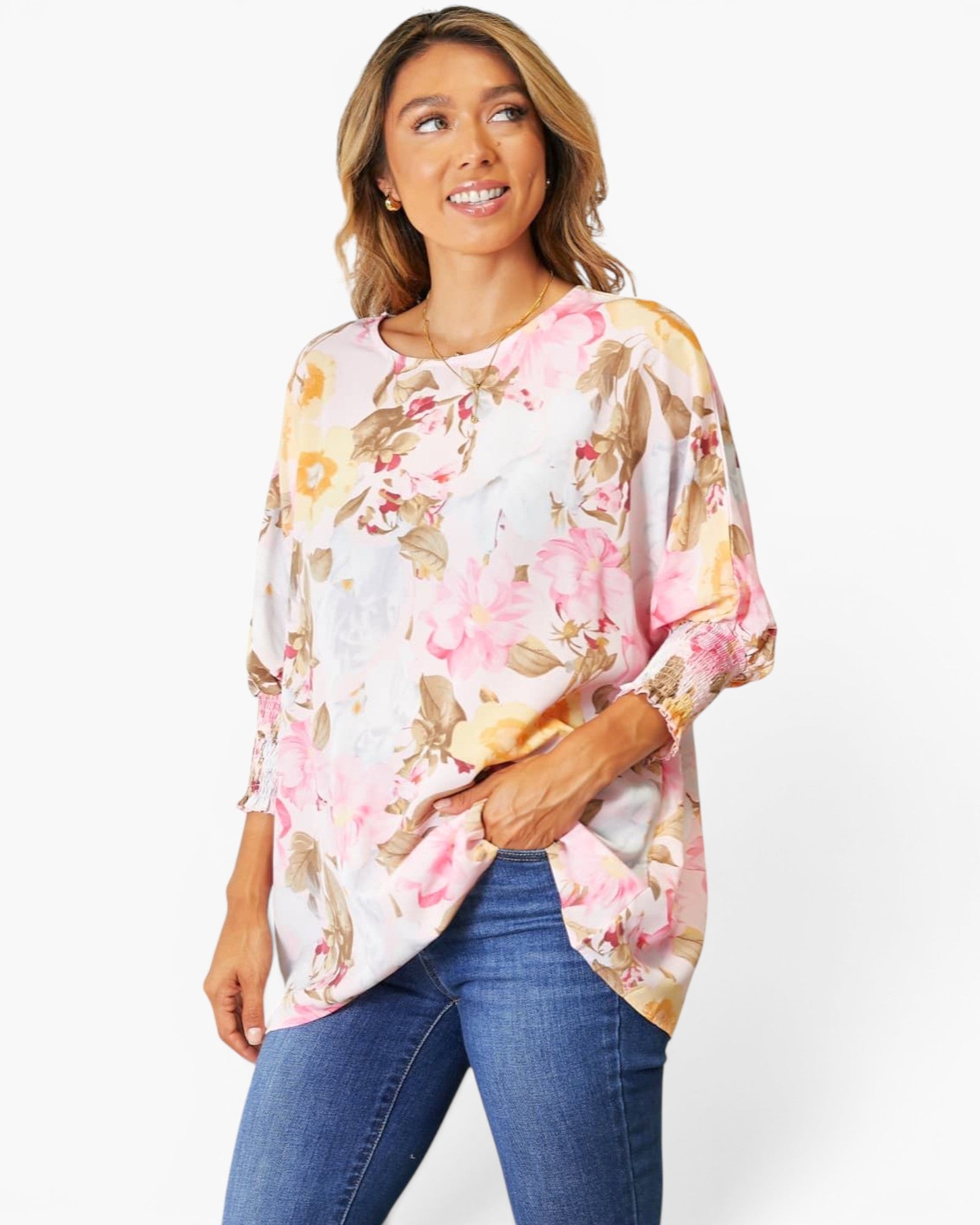 A woman wearing a loose, three-quarter sleeve floral top with pink, yellow, and beige tones over a white base, paired with blue jeans, against a plain white studio background.