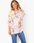 A woman wearing a white long-sleeve tunic top with a pink, yellow, and brown floral print, paired with blue jeans, against a plain white background.