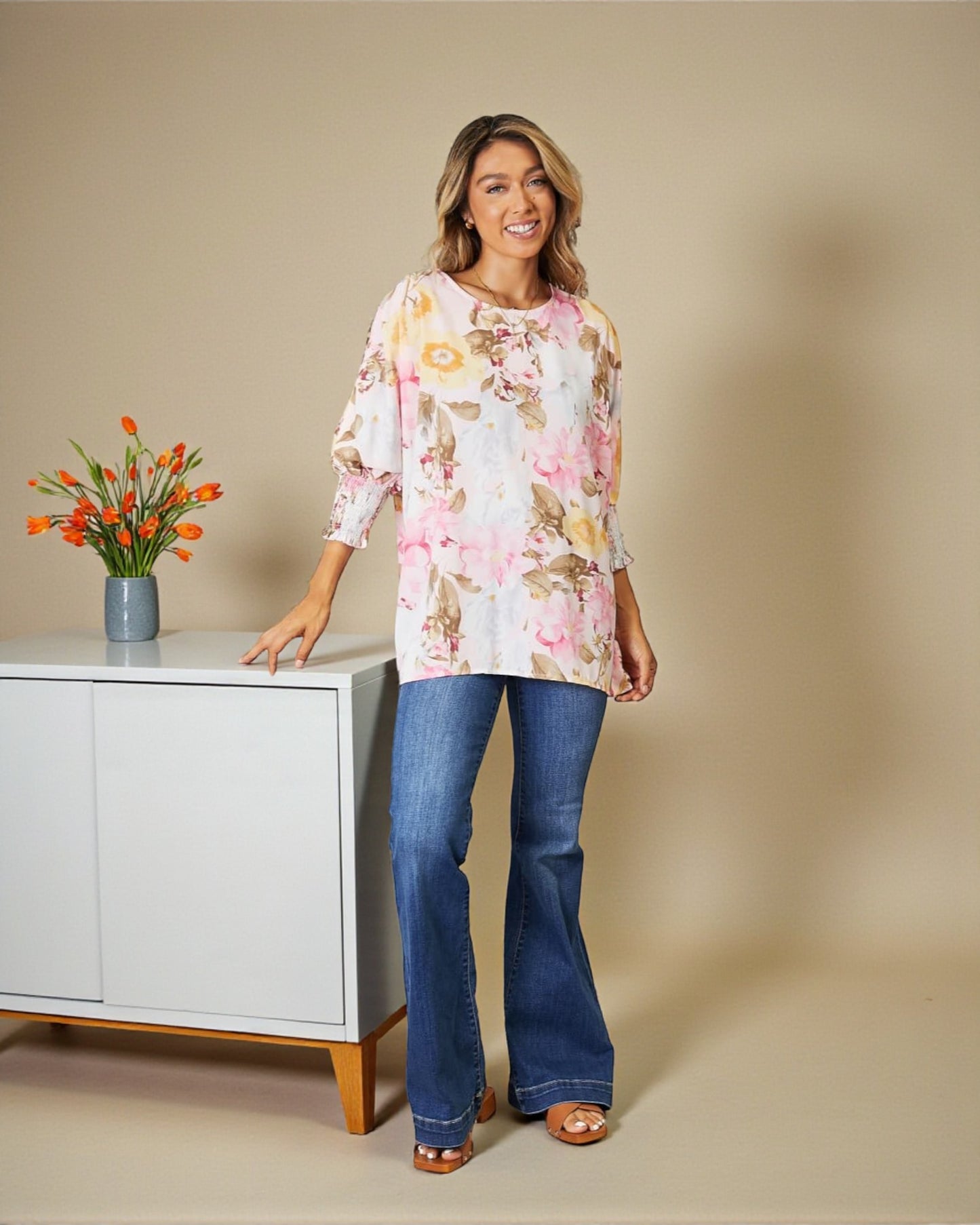 Woman wearing a floral blouse and blue jeans standing next to a white cabinet with flowers on it.