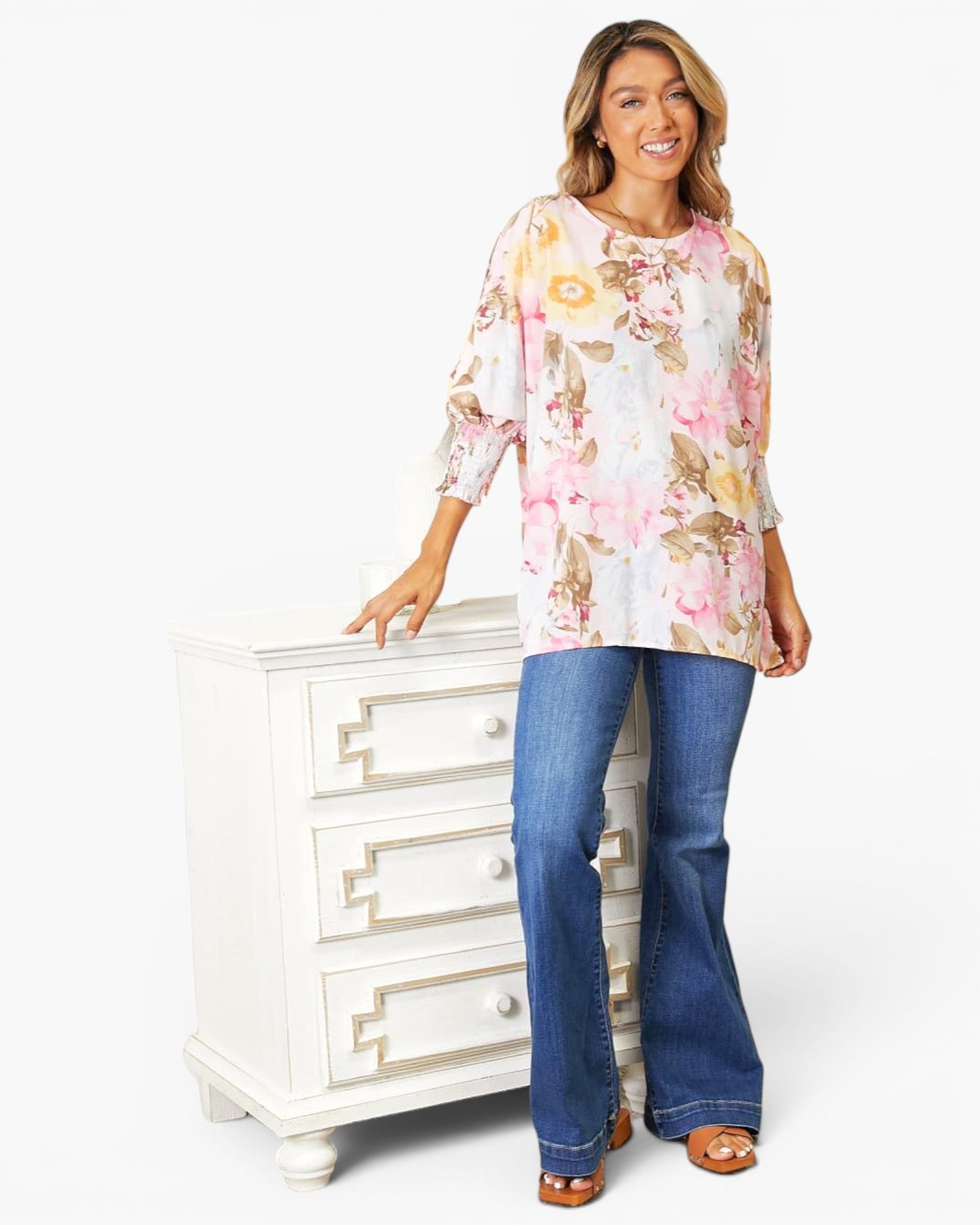 A woman modeling a three-quarter sleeve floral top with blue flare jeans, standing next to a white three-drawer chest against a white studio background.