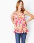 A woman modeling a sleeveless floral top with thin straps, featuring a vibrant pink, yellow, and white pattern, paired with blue jeans against a plain white background.