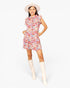 A woman modeling a short floral print dress with ruffled shoulders and pockets, paired with a wide-brimmed cream hat and tall cream boots, against a plain light gray studio background.