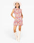 A woman modeling a short floral print dress with ruffled cap sleeves and a high neckline, paired with a wide-brimmed cream hat and cream knee-high boots, against a light gray studio background.