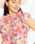 A close-up of a woman wearing a colorful floral print dress with ruffled shoulders and a tie neckline, paired with a light pink hat against a plain white background.