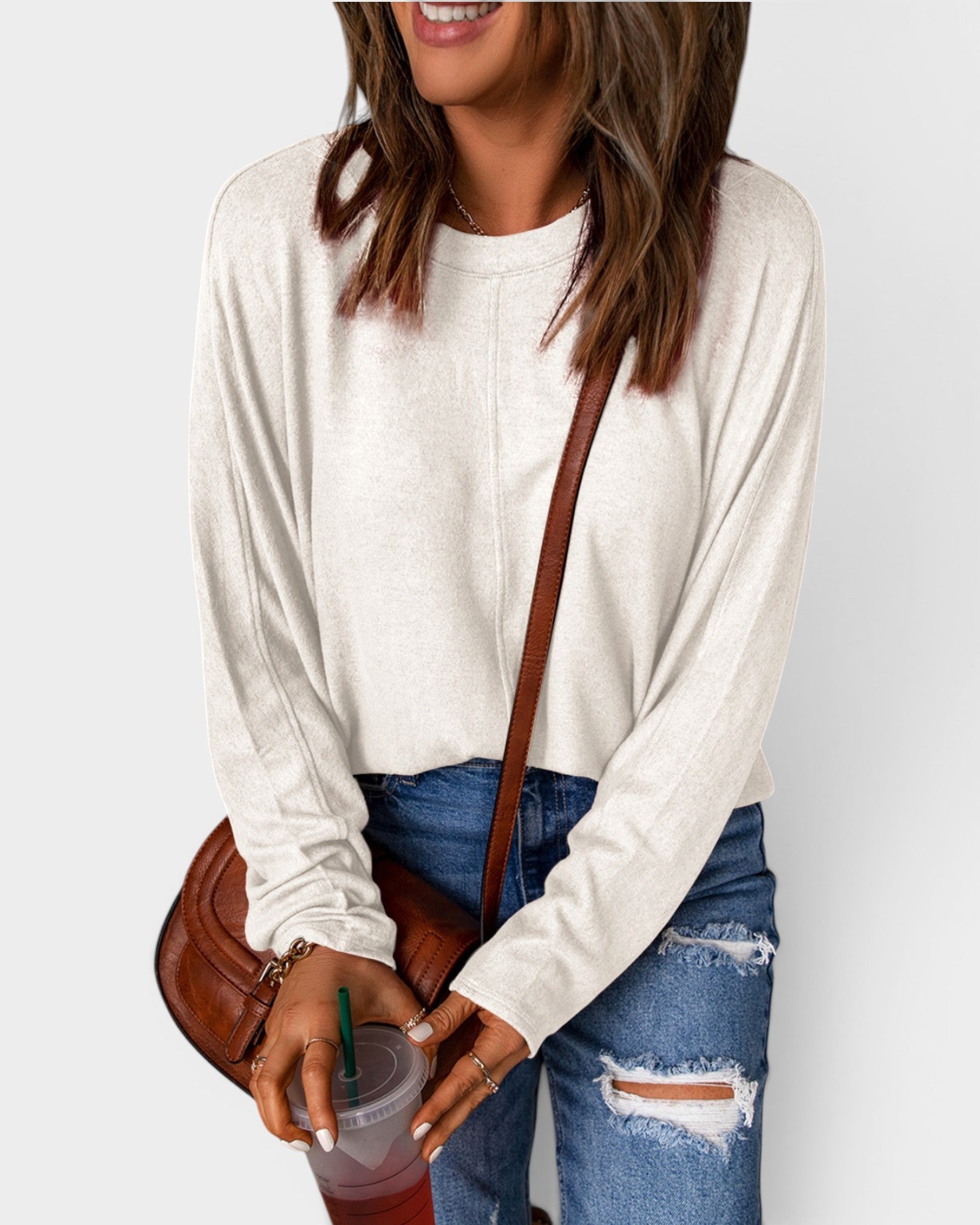 Person wearing a white long-sleeve top and blue jeans, holding a brown bag and a drink.