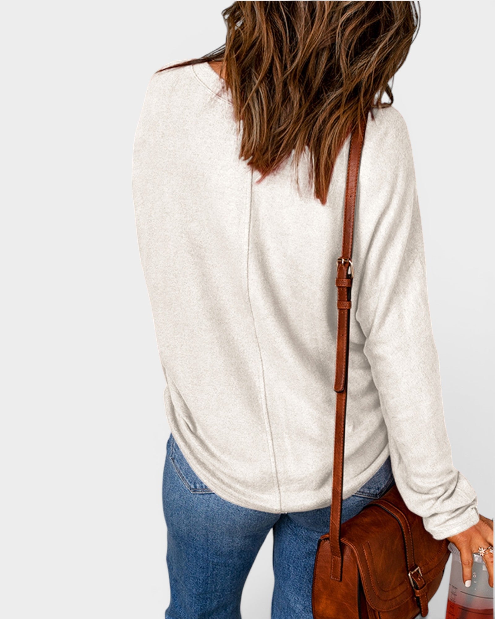Person wearing a white sweater and blue jeans with a brown leather bag on a light gray background