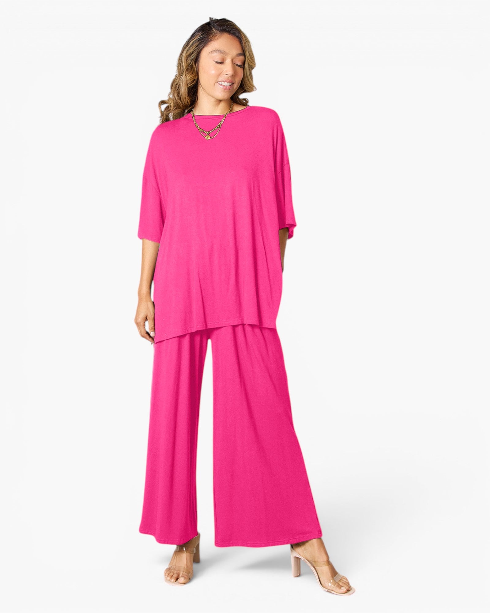 A woman modeling a bright pink two-piece lounge set consisting of a short-sleeve top and matching wide-leg pants, photographed against a clean white studio background.