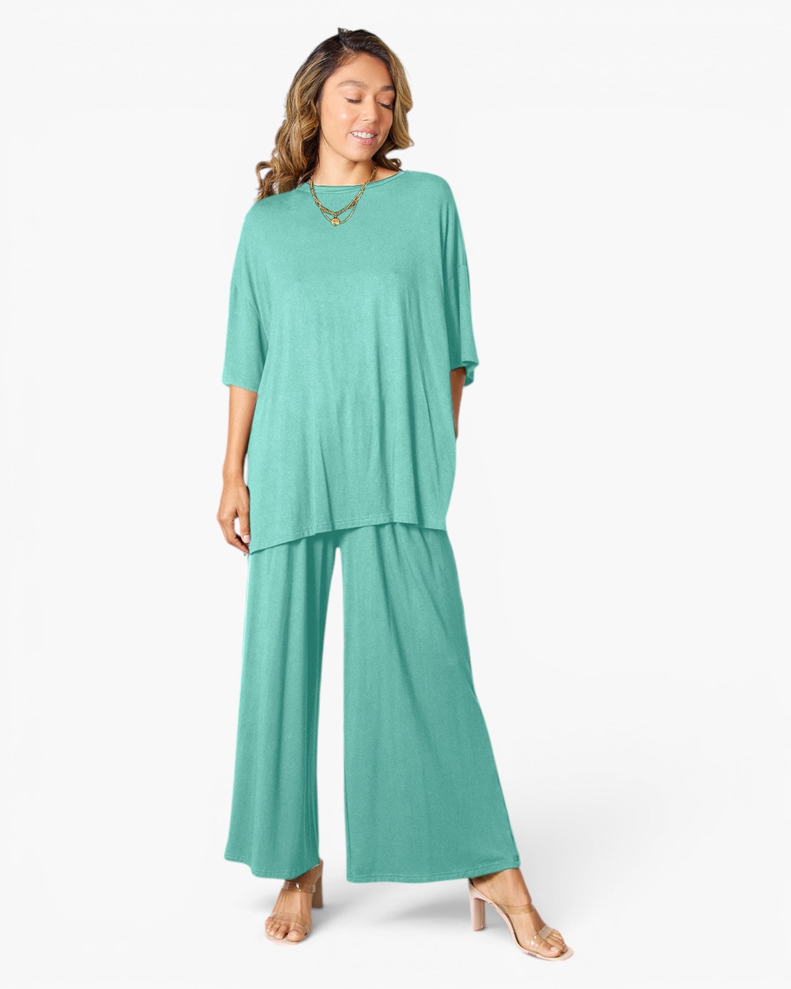 A woman modeling a matching two-piece outfit consisting of a short-sleeve, loose-fitting teal top and wide-leg teal pants, styled with a gold necklace and clear high-heeled sandals against a plain white studio background.