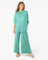 A woman modeling a matching two-piece outfit consisting of a short-sleeve, loose-fitting teal top and wide-leg teal pants, styled with a gold necklace and clear high-heeled sandals against a plain white studio background.