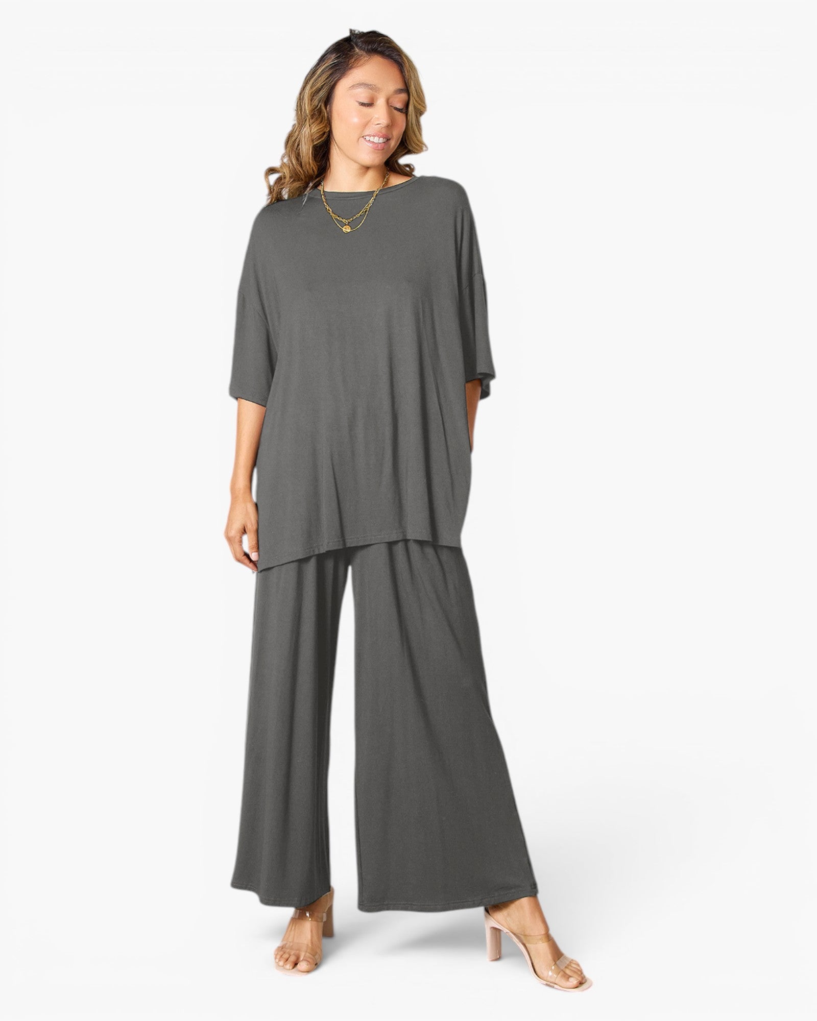 A woman modeling a dark gray two-piece lounge set consisting of a short-sleeve top and matching wide-leg pants, styled with gold necklaces and beige high-heeled sandals against a plain white background.