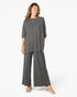 A woman modeling a dark gray two-piece lounge set consisting of a short-sleeve top and matching wide-leg pants, styled with gold necklaces and beige high-heeled sandals against a plain white background.