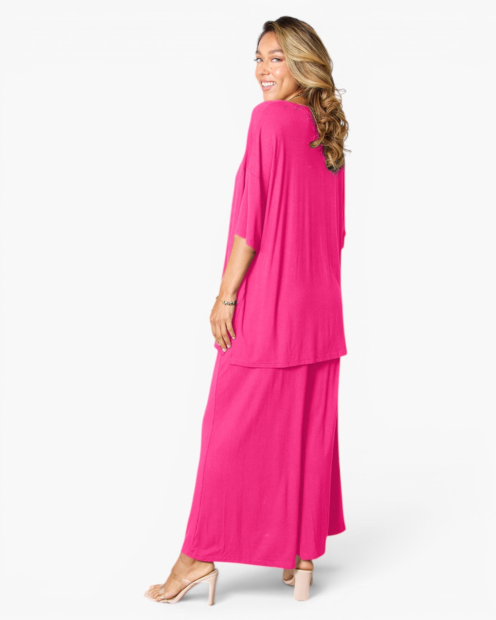 A woman modeling a bright pink two-piece outfit consisting of a short-sleeve draped top and matching wide-leg pants, shown from a side-back angle against a plain white studio background.