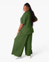 A woman modeling a matching olive green scrub set, featuring a short-sleeve top and wide-leg pants, shown from a three-quarters back view against a plain white background.