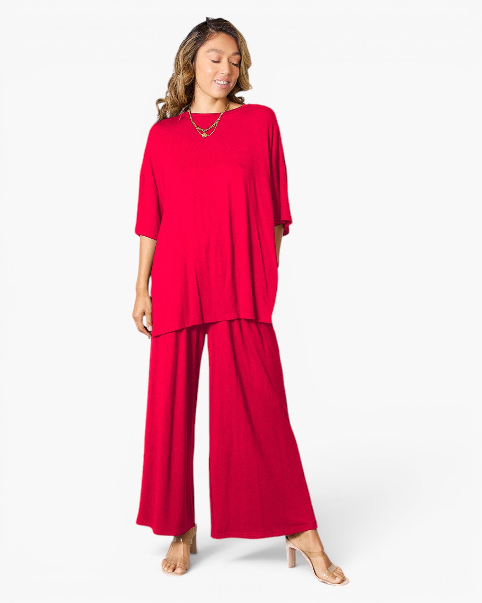 A woman modeling a bright red two-piece outfit consisting of a short-sleeve, loose-fitting top and matching wide-leg pants, paired with nude high-heeled sandals and layered gold necklaces, against a plain white studio background.