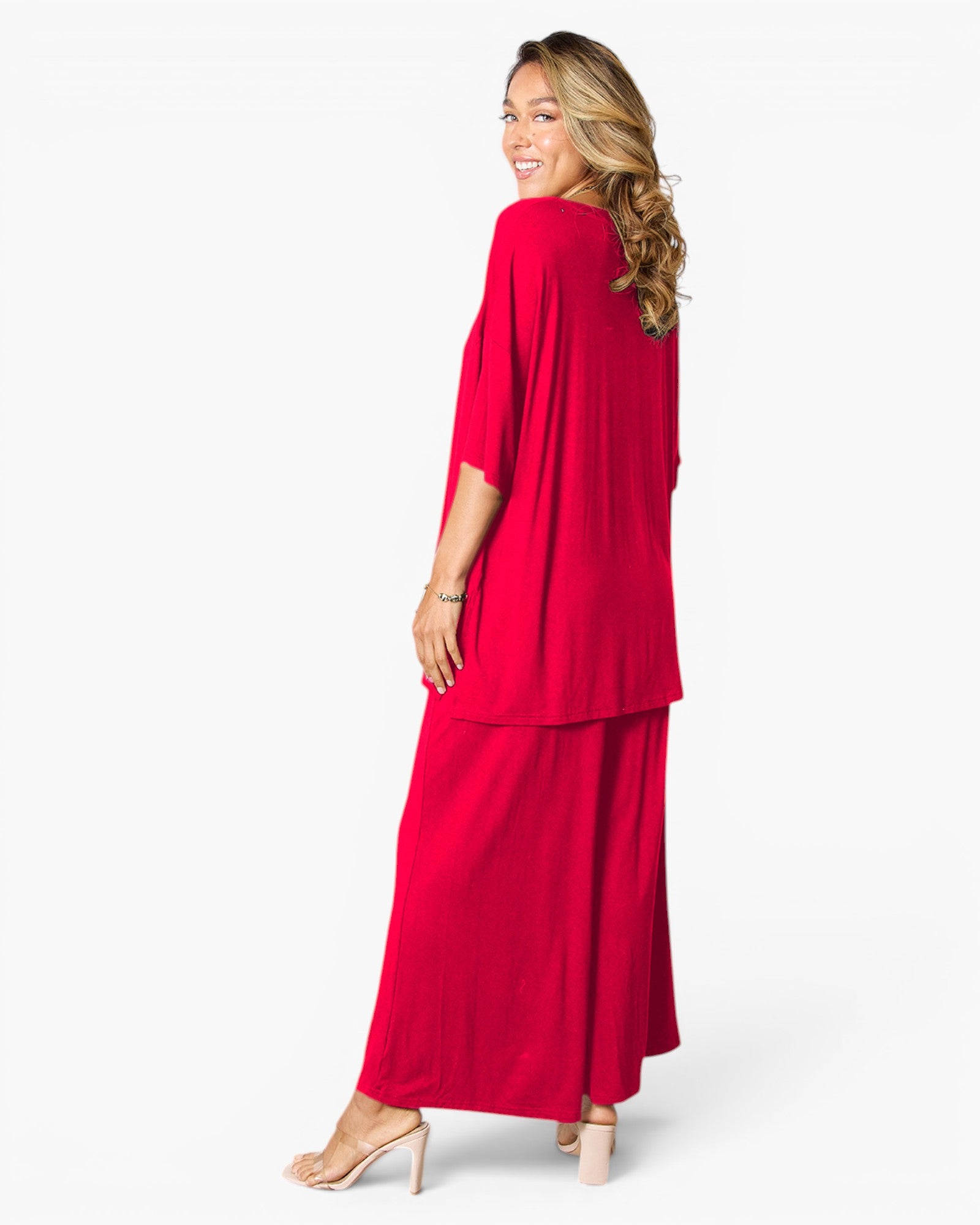 A woman modeling a bright red two-piece outfit consisting of a draped long-sleeve top and matching wide-leg pants, shown from a side-back angle against a plain white studio background.