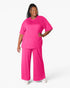 A woman modeling a bright pink two-piece lounge set consisting of a short-sleeve crewneck top and matching wide-leg pants, photographed against a clean white studio background.