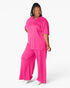 A woman modeling a bright pink two-piece loungewear set consisting of a short-sleeve V-neck top and matching wide-leg pants, photographed against a clean white studio background.