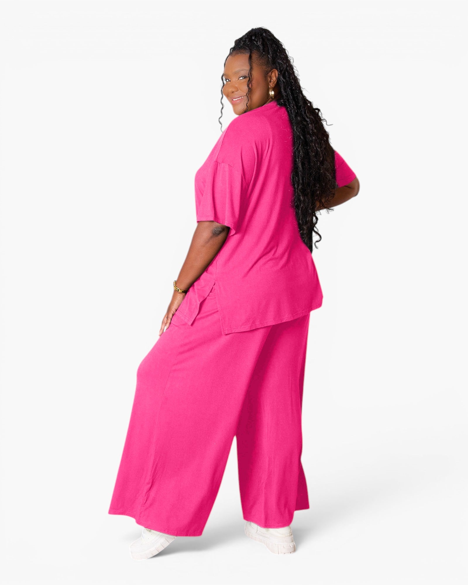 A woman modeling a bright pink scrub set consisting of a short-sleeve top and matching wide-leg pants, shown from a three-quarters back angle against a plain white studio background.