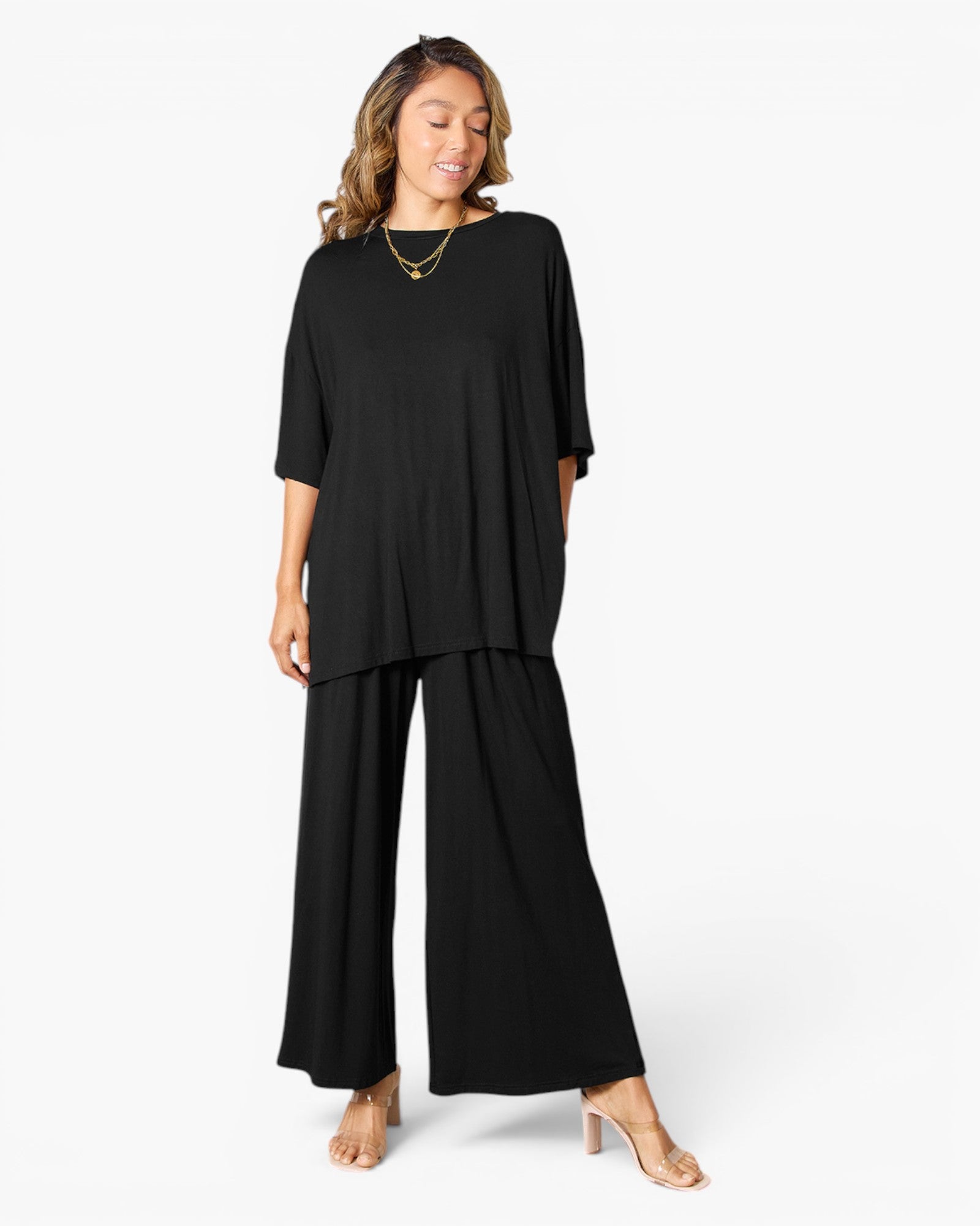 A woman modeling a black two-piece outfit consisting of a short-sleeve, loose-fitting top and matching wide-leg pants, styled with gold necklaces and beige high-heeled sandals against a plain white studio background.