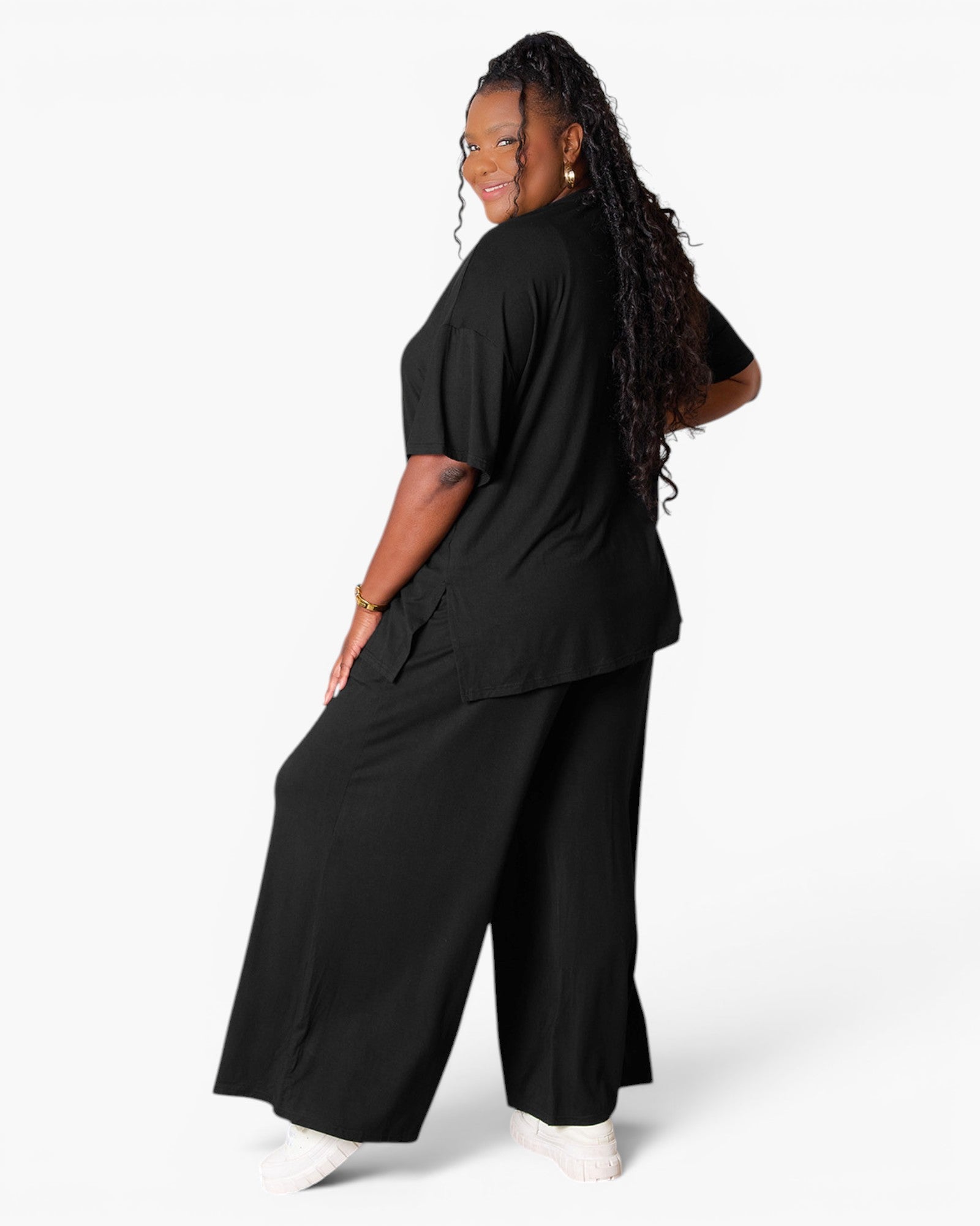 A woman modeling a black two-piece lounge set consisting of a short-sleeve top and wide-leg pants, shown from a three-quarters back view against a light gray studio background.