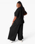 A woman modeling a black two-piece lounge set consisting of a short-sleeve top and wide-leg pants, shown from a three-quarters back view against a light gray studio background.