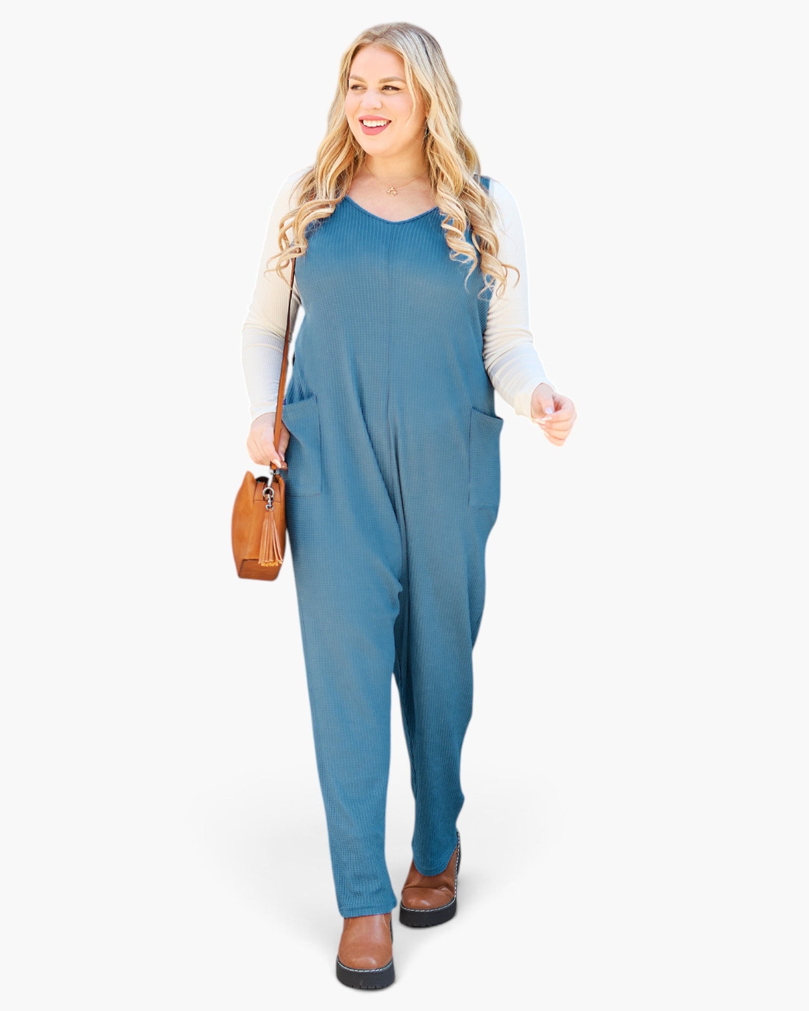 A woman modeling a slate blue sleeveless jumpsuit over a light beige long-sleeve shirt, holding a small brown leather handbag against a plain white background.