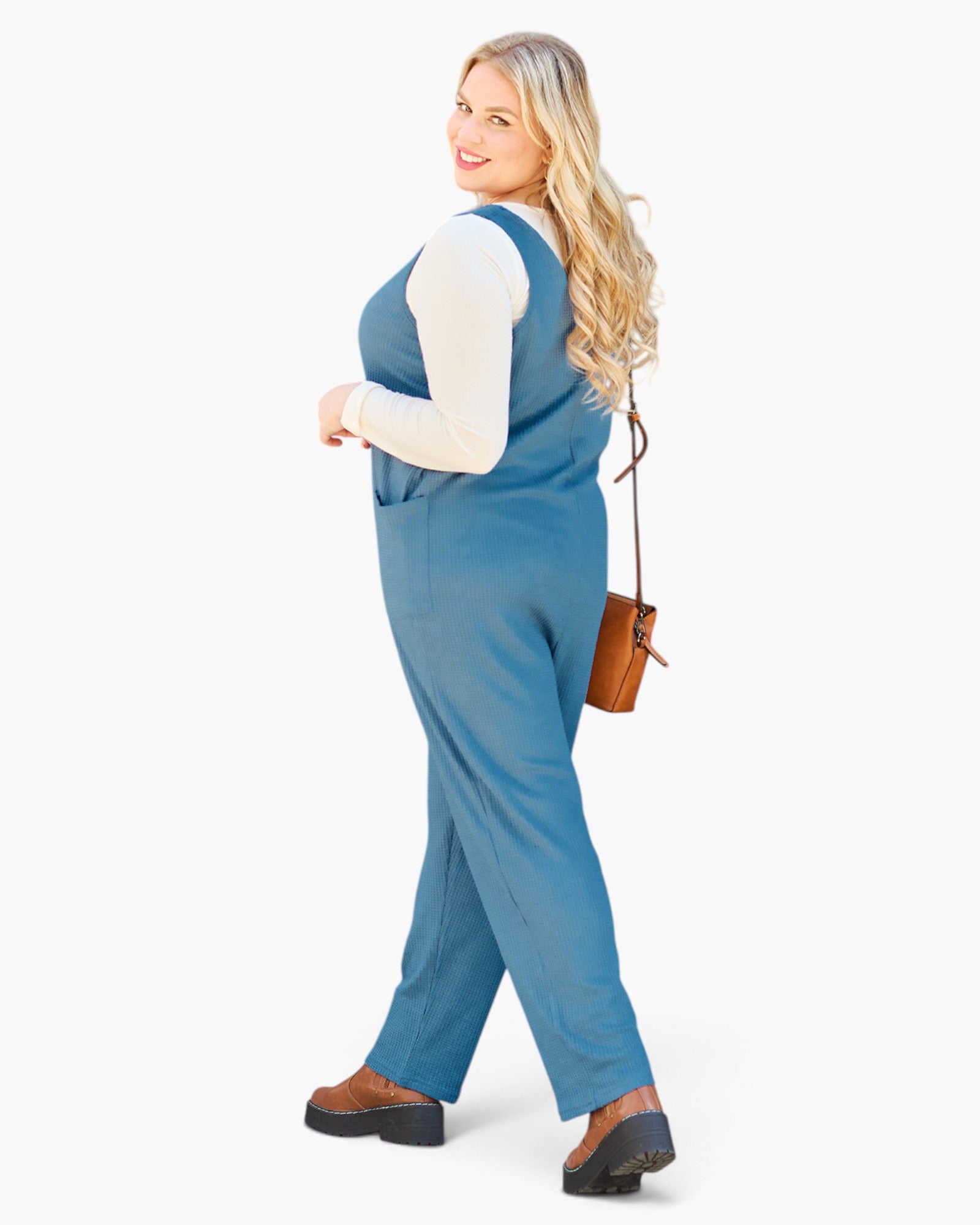 A woman modeling a slate blue sleeveless jumpsuit over a white long-sleeve top, wearing brown platform shoes and carrying a small brown shoulder bag, set against a plain white studio background.